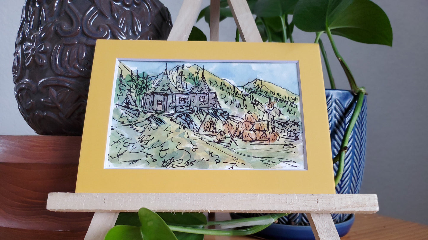 A framed ink and watercolor painting of Hagrid's hut in a pumpkin patch, displayed on a wooden easel.