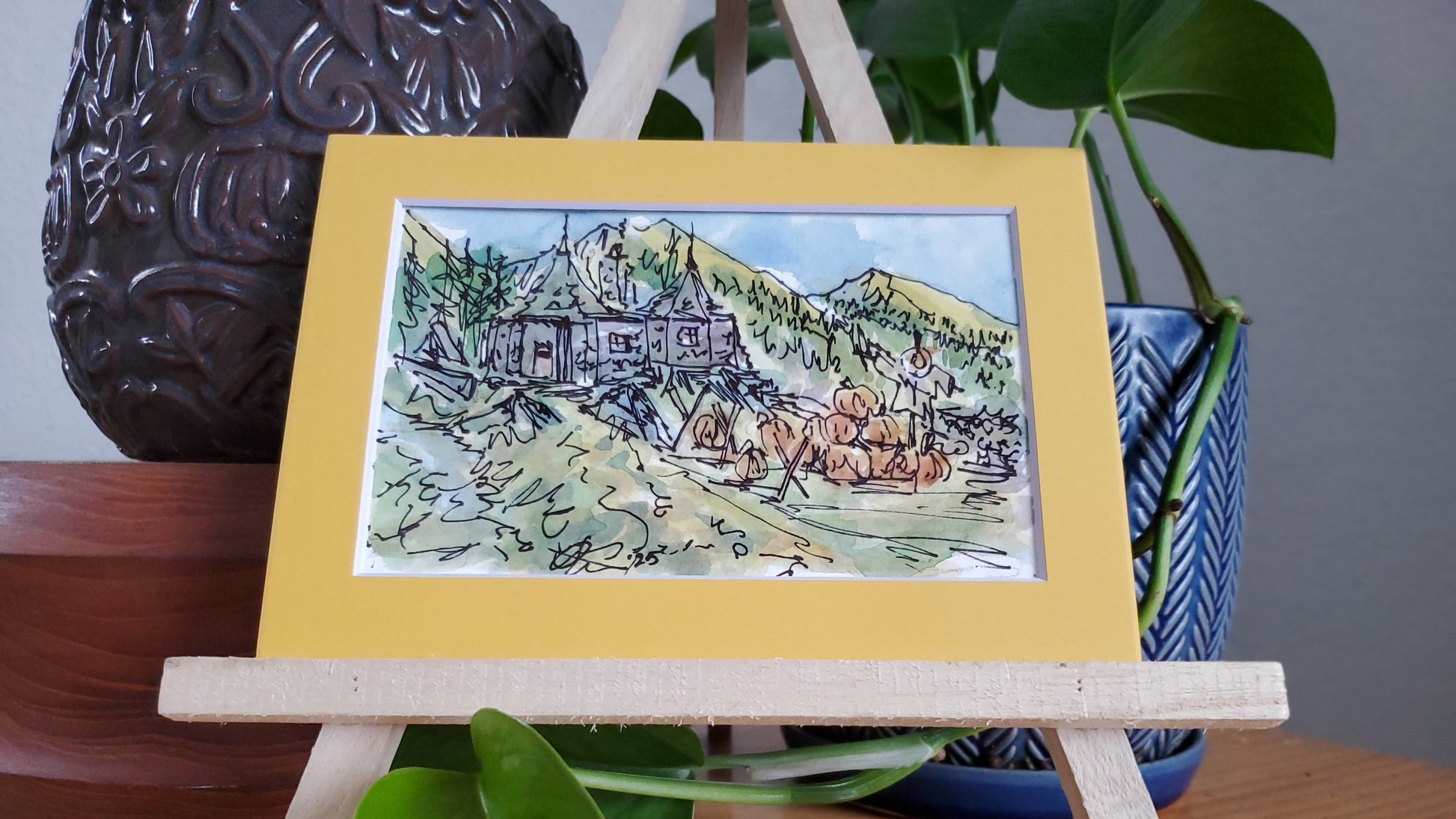 A framed ink and watercolor painting of Hagrid's hut in a pumpkin patch, displayed on a wooden easel.