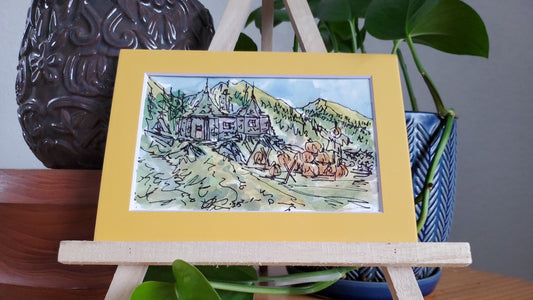 A framed ink and watercolor painting of Hagrid's hut in a pumpkin patch, displayed on a wooden easel.